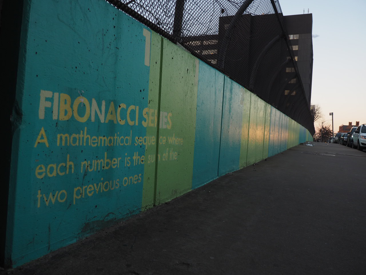 A mural depicting the Fibonacci series through a series of color blocks and the text "Fibonacci series: A mathematical sequence where each number is the sum of the two previous ones" on a wall along a sidewalk at sunset